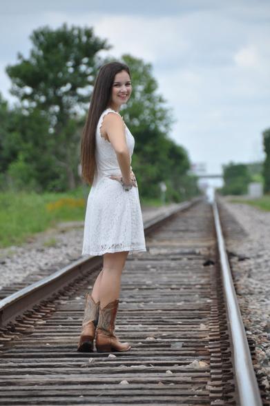 Emily Grice in white dress to her knee with cowboy boots. Long brunette hair. Standing in the middle of railroad tracks posing for graduation photos. Right hand on hip, looking right.