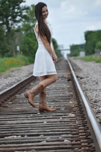 Emily Grice in white dress to her knee with cowboy boots. Long brunette hair. Standing in the middle of railroad tracks posing for graduation photos. Looking right, right foot in the air.