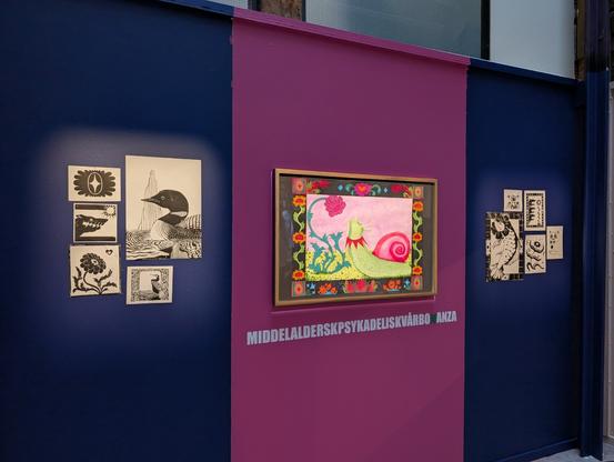 Picture of a wall with art on it. Its painted dark blue with a big pink stripe in the middle. On the pink section there's a huge drawing of a snail and lots of colorful flowers. On both of the sides on the blue parts there's some clusters of lino prints of flowers, birds, fish and other fun stuff.