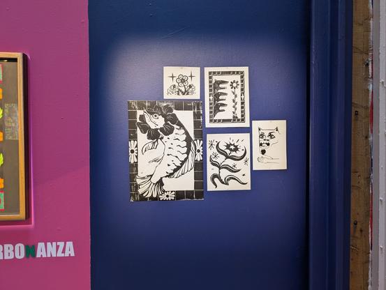 Close up picture of a section of the wall. Its painted blue and hanging on the wall there's lino prints of a fish with a flower around it's head, two flowers, a cat man creature with cool facial hair and one of some dog creatures screaming at a flower