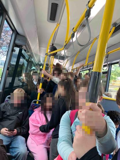 Viele Schüler:innen im Linienbus. Auch alle Stehplätze belegt. Bild ist in Farbe. Rosa und hellblaue Kleidung zweier Schüler:innen fallen auf. Gesichter verpixelt. Haltestangen sind gelb.