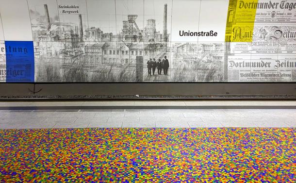 A subway platform with a colorful tiled floor and a wall with a mural of older industrial buildings and old newspaper clippings.

Ein U-Bahn-Bahnsteig mit buntem Kachelboden und einer Wand mit einer Wandmalerei älterer Industriegebäude und alten Zeitungsausschnitten.