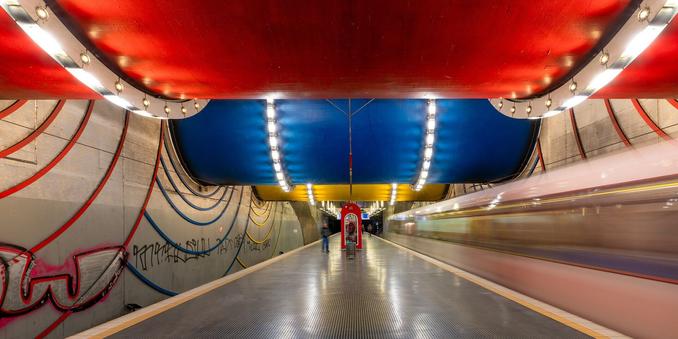 U-Bahnhof mit Mittelbahnsteig. Auf dem rechten Gleis fährt ein Zug ein. An der Decke befinden sich drei große Röhren, in den Farben Rot, Blau und Gelb, mit Lichtern verziert.