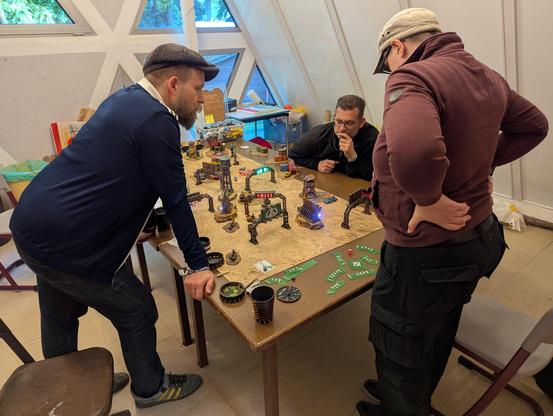 In der Mitte eines Raumes steht ein Tisch auf dem ein Tabletop-Spiel aufgebaut ist. Die Dekoration ist aus einer apokalyptischen Welt. Statt Figuren stehen Autos wie aus Mad Max auf dem Feld verteilt. Um den Tisch stehen zwei Personen, eine dritte sitzt. Alle starren wie gespannt auf das Feld.