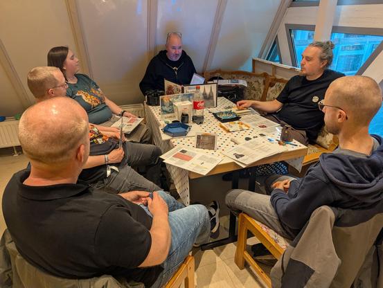 Eine Gruppe von Personen sitzt um einen Tisch herum. Auf dem Tisch verteilt liegen Blätter, Würfel und Getränke. Am Kopfende sitzt der Spielleiter, der aus einem Buch vorliest. Die Spieler schauen ihn erwartungsvoll an.