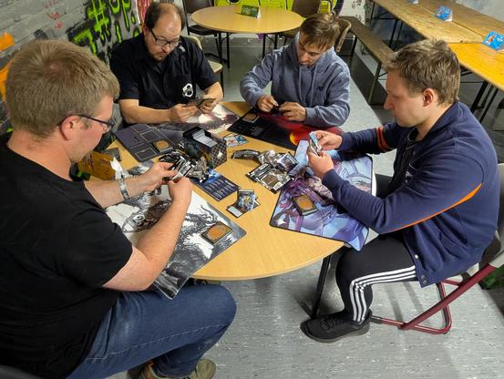Vier Spieler sitzen um einen runden Tisch herum. Jeder von ihnen hat eine Spielmatte vor sich liegen und hält Karten in den Händen. In der Tischmitte liegen mehr Karten. Alle sind in ihre Karten vertieft.