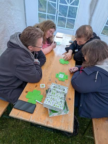 An einem Tisch sitzen vier Personen. Sie schauen alle auf eine Art vierblättriges Kleeblatt aus Kunststoff. Eine der Personen zeigt den Spielern etwas darauf.