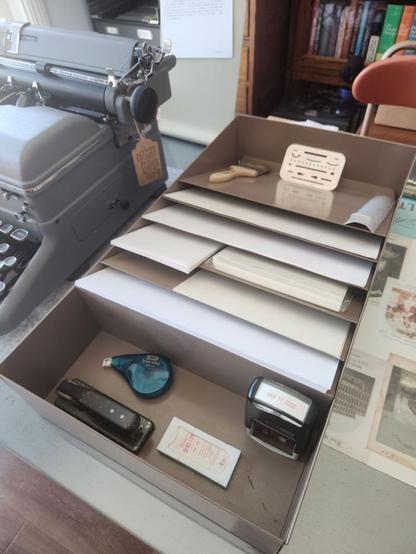 Angle down from just above a paper organizer. We can see a variety of blank paper types and sizes filed into the angled slots . In the very back we see a paint chip brush for dusting a typewriter as well as a couple different eraser shields for typing and error correction.