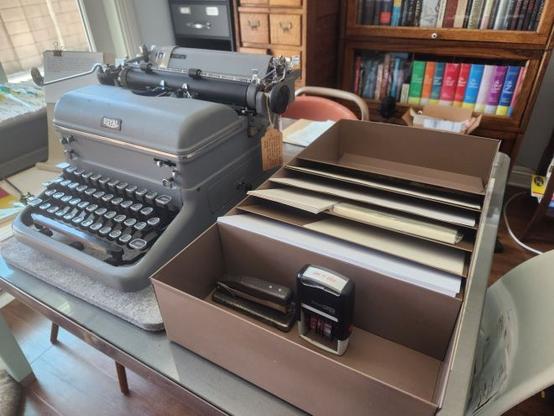 Oblique view of a Hunt Manufacturing paper organizer in 20 gauge steel next to a Gray Royal KMG typewriter. The drawer organizer has six angled slots for storing and organizing 8.5 x 11" pieces of paper. In the front of the organizer is a 4.5 x 11.5 inch section for various office and typing supplies. In this section we see a stapler and a rubber date stamp.