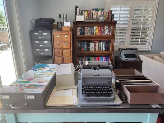 Analog office set up featuring a stick leg metal desk, a Royal KMG typewriter, a few card index filing cabinets, a typing copy stand, and a paper supply organizer.