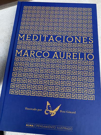 Una copia de Meditaciones de Marco Aurelio apoyado en el alféizar de una ventana. Es una portada azul con decoraciones geométricas amarillas que recuerdan un mosaico romano entre las que figuran el título y autor de la obra.