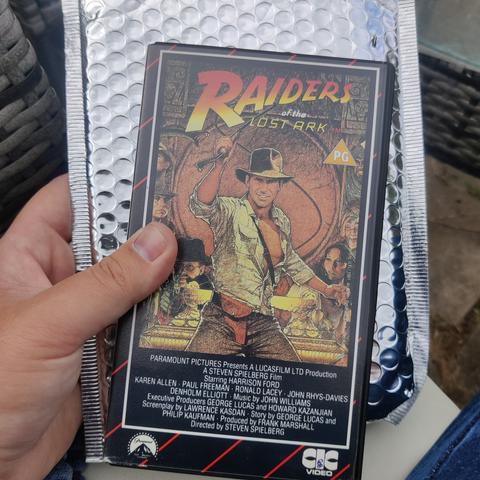 A VHS tape of Raiders Of The Lost Ark held up with its shiny silver postage packaging as a background. The box features the classic film poster but with a stripy red and black background around the edges.