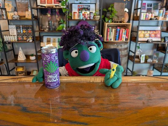 A green puppet with purple hair & nose is sitting at a bar in a weed store. They are holding a can of THC juice and a yellow chillum.