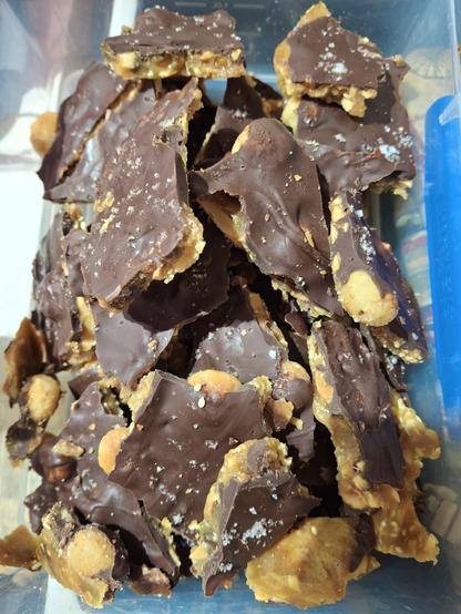 A box of broken pieces of chocolate topped, caramel brittle. Some of the seeds and nuts, particularly the large macadamias can be seen in the caramel layer. The chocolate topping is sprinkled with sea salt.