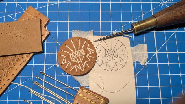 Auf einer Schneidmatte liegt eine Vorlage aus Papier mit Resten von Klebeband, die eine stilisierte Distelblume als Strichzeichnung zeigt. Die Schnittpunkte der Linien sind mit einer spitzen Ahle auf ein rundes Stück cognac-braunes Leder übertragen worden, das daneben liegt, und durch die entstandenen Löcher ein im Vergleich relativ dicker, weißer Faden gezogen wurde. Im äußeren Bereich des Bildes liegen noch ein Heftchen Ledernadeln mit stumpfer Spitze, die erwähnte Ahle, und ein paar Reststücke, die beim Durchstechen als zusätzliche Unterlage gedient haben.