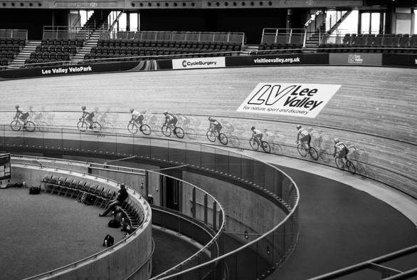 A black and white photograph shows an indoor cycle track, the Lee Valley VeloPark, with a group of cyclists racing. The track curves steeply, and the cyclists are captured with a motion blur effect, creating a sense of speed. Banners with "Cycle Surgery" and "visitleevalley.org.uk" are visible along the track's edge. In the background, there are rows of empty seats. The overall scene captures the dynamic energy of a cycling event in a modern velodrome.