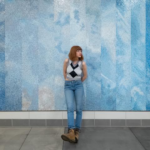 Photo of Lety standing in front of a blue mosaic mural under white LED lighting at an underground train station. From top to bottom, she is shown wearing a plaid argyle cropped sweater vest, blue jeans, and brown boots. Lety is centered to the photo, her hands clasped behind her butt and her legs angled out in front of her as she leans on the mural “At the Same Time” by Rebeca Méndez. She's trying to look super cool and casual, her head facing toward image-right as if she were looking for an oncoming train, even though she's literally an entire floor above the actual train stop.