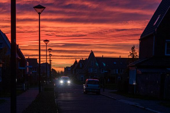 In the middle of the photo you see a street with sidewalks on both sides and rows of houses. It is almost dark but you can just make out the houses. The windows and white parts of the houses light up white or red because of the setting sun. The sky is incredibly orange with wisps of clouds in between. On the street in the middle there is a parked car on the right. The roof is red because of the sun. A car is approaching on the left, with its lights on. Along the sidewalk on the left is a long row of lampposts. The glass lamps on the poles are red because of the light that shines through them. There are lines from left and right towards the middle, from the curbs, the street and the lamps.