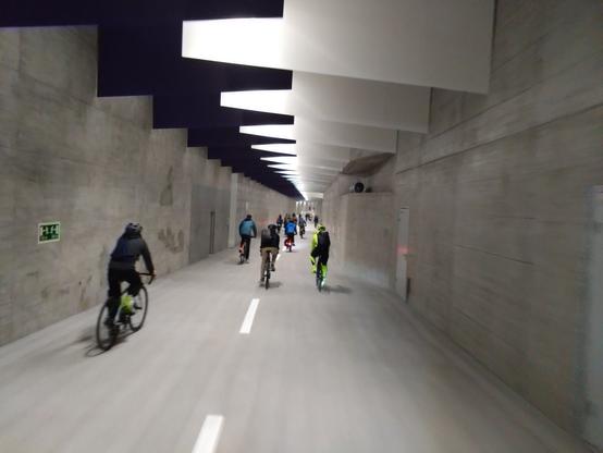Ein Foto vom neuen Velotunnel, man sieht etwa 100 m weit bis zu hinteren Rampe, bis dort hat es etwa 20 Menschen auf ihren Velos. An der Decke hängen farbige Panele.