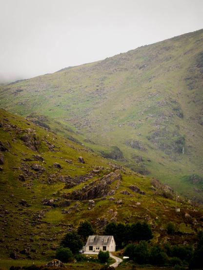 An eery photo of a white house at the bottom of a misty, rocky, green valley.