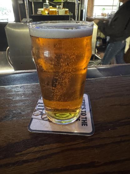 A pint of amber beer sits on a wooden bar top, resting on a coaster. In the background, blurred figures and bar elements are visible, suggesting a lively atmosphere.