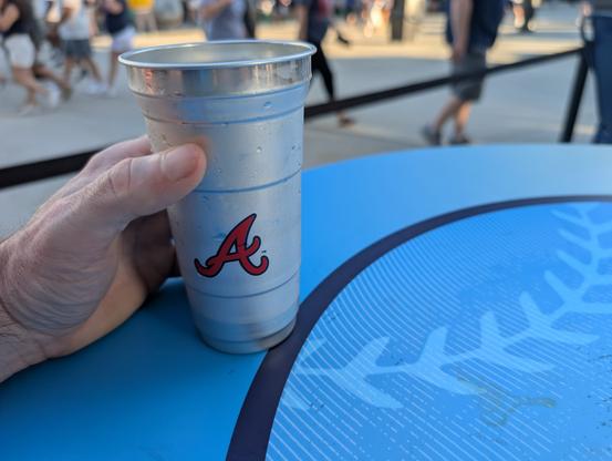 The image shows a hand holding a silver plastic cup with a red "A" logo on it, which is likely associated with the Atlanta Braves baseball team. The cup is resting on a blue table with a circular design featuring a white antler-like pattern. The background is blurred, but it appears to be an outdoor setting with people walking around, suggesting a public event or gathering. The lighting indicates it is daytime, and the overall atmosphere seems casual and relaxed.