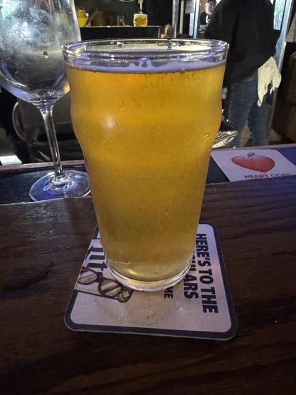 A glass of beer fills the foreground, showcasing a golden hue and a frothy top. In the background, there's a blurred empty wine glass and hints of a bustling bar scene. The beer is placed on a coaster with text visible.