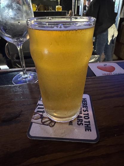 A glass of beer fills the foreground, showcasing a golden hue and a frothy top. In the background, there&#039;s a blurred empty wine glass and hints of a bustling bar scene. The beer is placed on a coaster with text visible.