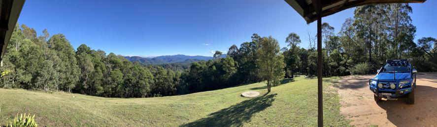 A panoramic view of the Brogo River valley and Wadbilliga National Park in the distance. Heavily forested hills precede similarly rugged mountains with a deep azure sky above. A low cloud bank is visible against the distant mountain peaks.