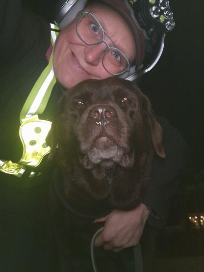 Night time selfie of a person and a chocolate brown Labrador with greying snout. The person is wearing a black jacket, yellow safety harness, red cap, silver headphones and blue-rimmed glasses. On their cap rests a headstrap torch in off position. Their free hand is around the dog's shoulders. Both are facing the camera. The person's head is resting on the dog's head. They're smiling, grateful for their time spent together.