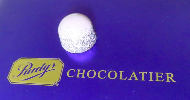 Lavender-colored bonbon with sides that have edible glitter. It's posed on top of the package it came in, a rich violet foil box with gold foil text. "Purdy's Chocolatier".