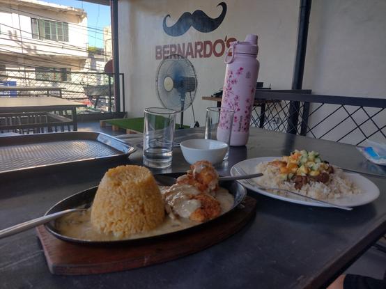 Table in a small restaurant featuring Sizzling Chicken and Pork Budbod. On the wall is logo for the restaurant "Bernardo's"