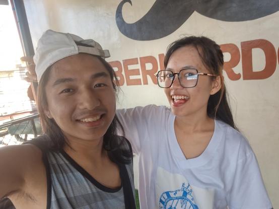 Selfie of a couple in front of is restuarant's logo ("Bernardo's" under a mustache logo) on wall