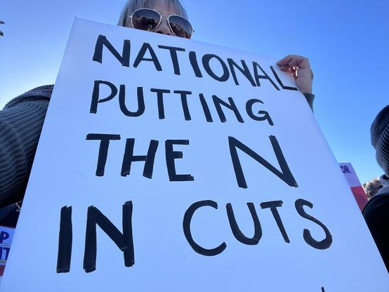 Image shows a person in sunglasses holding up a hand painted sign saying 

NATIONAL 
PUTTING
THE N 
IN CUTS