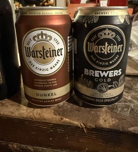 Two cans of beer sitting on a beat up workbench. The left is light brown and gold, the right is black and gold