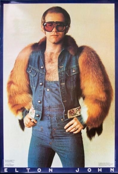 Elton John wearing a denim jacket with fur sleeves over tight denim overalls, hands on hips and large, subdued for Elton, sunglasses.