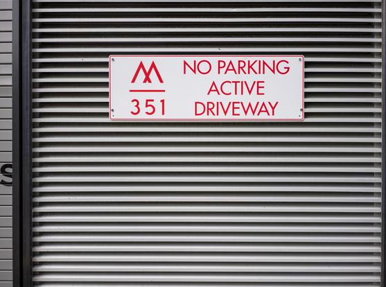 A white sign reading NO PARKING / ACTIVE / DRIVEWAY (along with a stylized "M" and the number 351) in red lettering is attached atop a wall section of horizontal louvers, which form a strict geometric pattern.  There's a bit of metal frame visible on the left, as well as the stenciled letter "S".