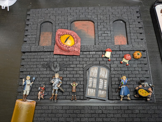 A miniature dungeon wall and floor primed in black, with most of the painted bits sitting on it. This includes a painted red dragon eye, a wooden door, 2 walking mushrooms, a dryad pumpkin, and 6 main characters from Delicious in Dungeon