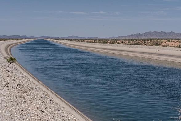 A color landscape photo of a large wide aqueduct filled with deep blue water flowing in a concrete channel from the lower right to the left center of the frame. A desert landscape is visible on the right side of the aqueduct. A distant mountain range is seen on the right as well.