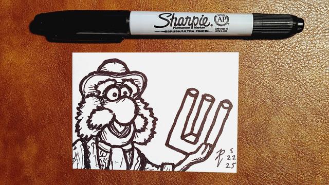 Marker drawing of Uncle Traveling Matt Fraggle proudly holding up an object which is the optical illusion known as a "poiuyt" or an "impossible trident," a shape which appears to have either two prongs or three. The drawing is signed with my "RV" signature glyph and dated 5-22-25. The black marker used to draw the image rests on the table above the card.