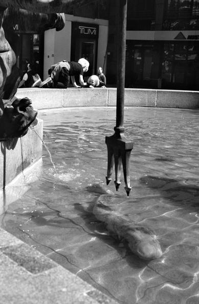The black and white image shows a fountain with a trident sculpture above water, a fish statue submerged below, and two children leaning over the edge, peering into the water. A building with shopfronts forms the background.