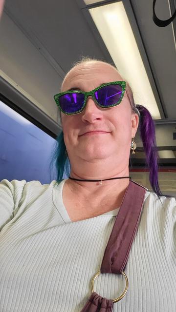 A selfie of me in a lime green top with sparkly green glasses, the roof and windows of a light rail car in the background