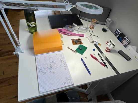 My living room table, white, folds out from a side table, also white. Scattered upon it are electronic socket components, green dot boards, an old brown dot board prototype, a steel rule, a pink, a blue, and a black erasable pen, an orange-covered gridbook with a complex partial wiring layout diagram drawn on the white page, a pink lanyard attached to another contraption with glasses rigged to it, and a few other everyday table items. A spring-armed table lamp magnifier is clamped to one side and gives extra light.