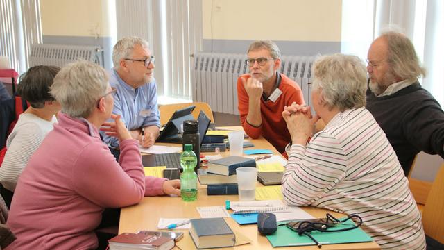 Um einen Tisch sitzen drei Frauen und drei Männer. Sie sehen aufmerksam auf einen der Männer. Er spricht gerade. Auf dem Tisch befinden sich ein Laptop, ein Tablet-PC, Mobiltelefone, Gesangbücher, bedrucktes Papier, Schreibutensilien und Getränke.