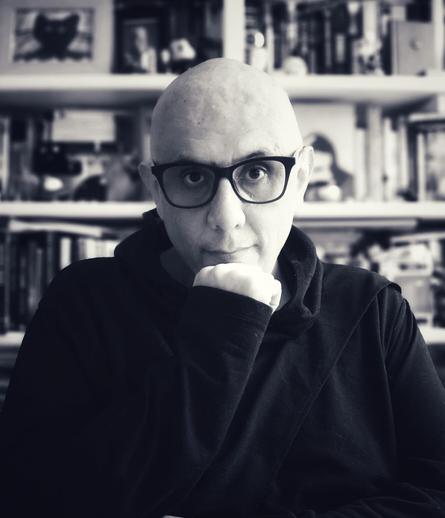 Un hombre de mediana edad, vestido de negro, con lentes de armazón negro y la cabeza afeitada, posa con una mano en la barbilla delante de un estante repleto de libros y objetos.