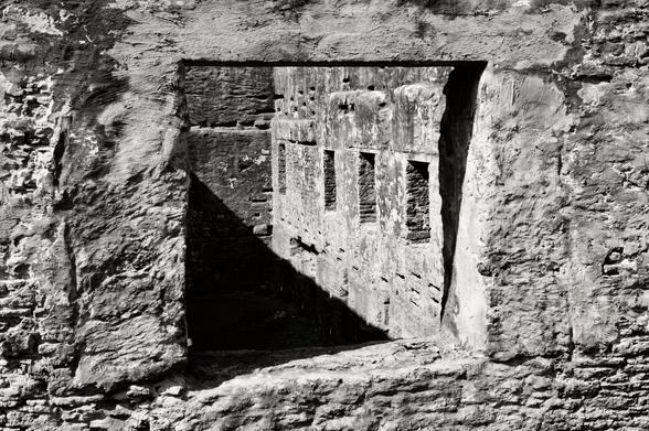 Black and white image of an old stone building.  The roof is gone so daylight streams inside from above.  The view is looking from outside through a window opening. An inner wall with more windows is visible, as well as a shadow cast across the inner wall.