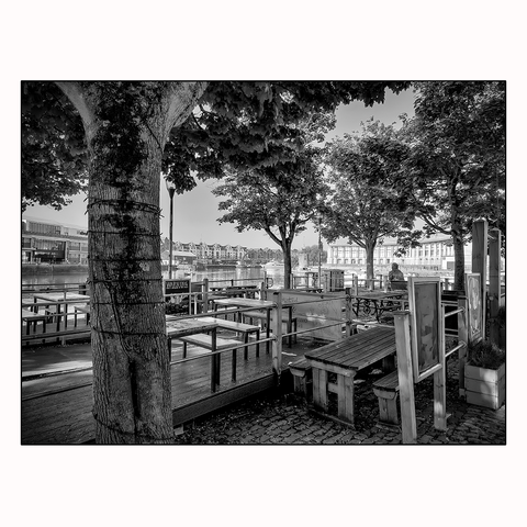 A quiet city scene overlooking the harbour in the distance with tables and chairs surrounded by trees that have mini lights wrapped around them.
