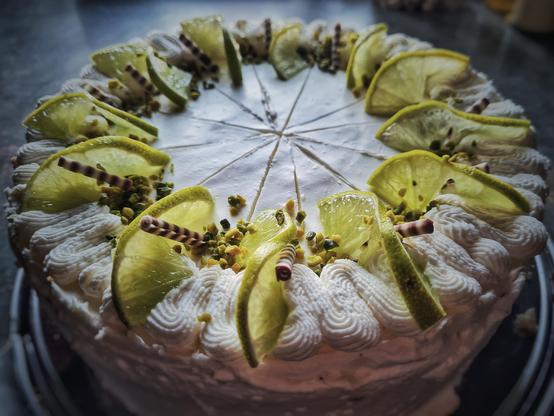 Das Bild zeigt eine kunstvoll dekorierte Kuchenplatte. Der Kuchen hat eine glatte, weiße Schicht mit einer kunstvollen Rüschenkremearbeit am Rand. Um den Rand herum sind frische Zitronenscheiben in gleichmäßigen Abständen angeordnet, die einen frischen und leuchtenden Kontrast zur weißen Schicht bilden. Zwischen den Zitronenscheiben sind kleine, braune und weiße Schokoladenstäbchen sowie grüne Pistazienstückchen verteilt, die dem Kuchen eine zusätzliche Textur und Farbe verleihen. Der Kuchen ist in gleichmäßige Stücke geschnitten, was auf eine sorgfältige Vorbereitung hinweist.