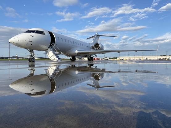 Bei weissblauem Himmel steht ein großer, weißer, zweistrahlige Privatjet (Global Express) mit geöffneter Eingangstür auf dem Rollfeld. Dieses ist vollständig mit Regenwasser bedeckt, wodurch sich die gesamte Maschine am Boden spiegelt. Die Aufnahme ist von sehr tief unten gemacht.