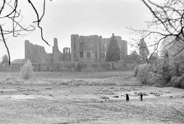 Black and white low contrast image of a ruined castle seen across a frost-covered field. A well-wrapped couple and their dog are playing on an area of ice. Twigs from a bush frame the image from above.
